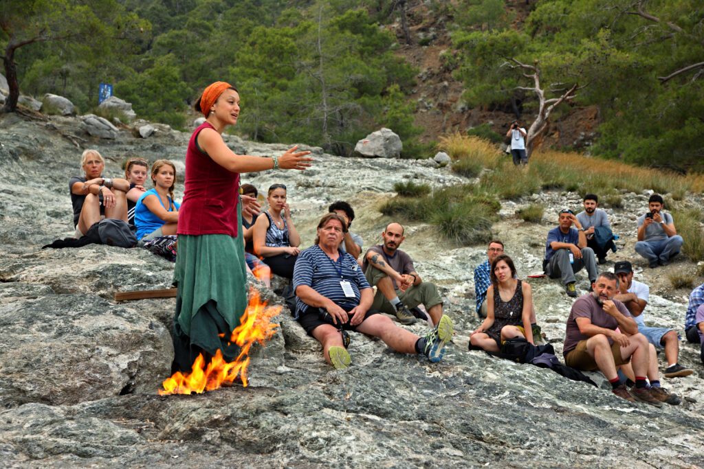 A woman by a campfire standing and telling a story to a group of people sitting on rocks