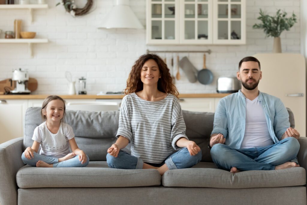 A mom and dad meditate on couch with small child.  This can develop social responsibility.