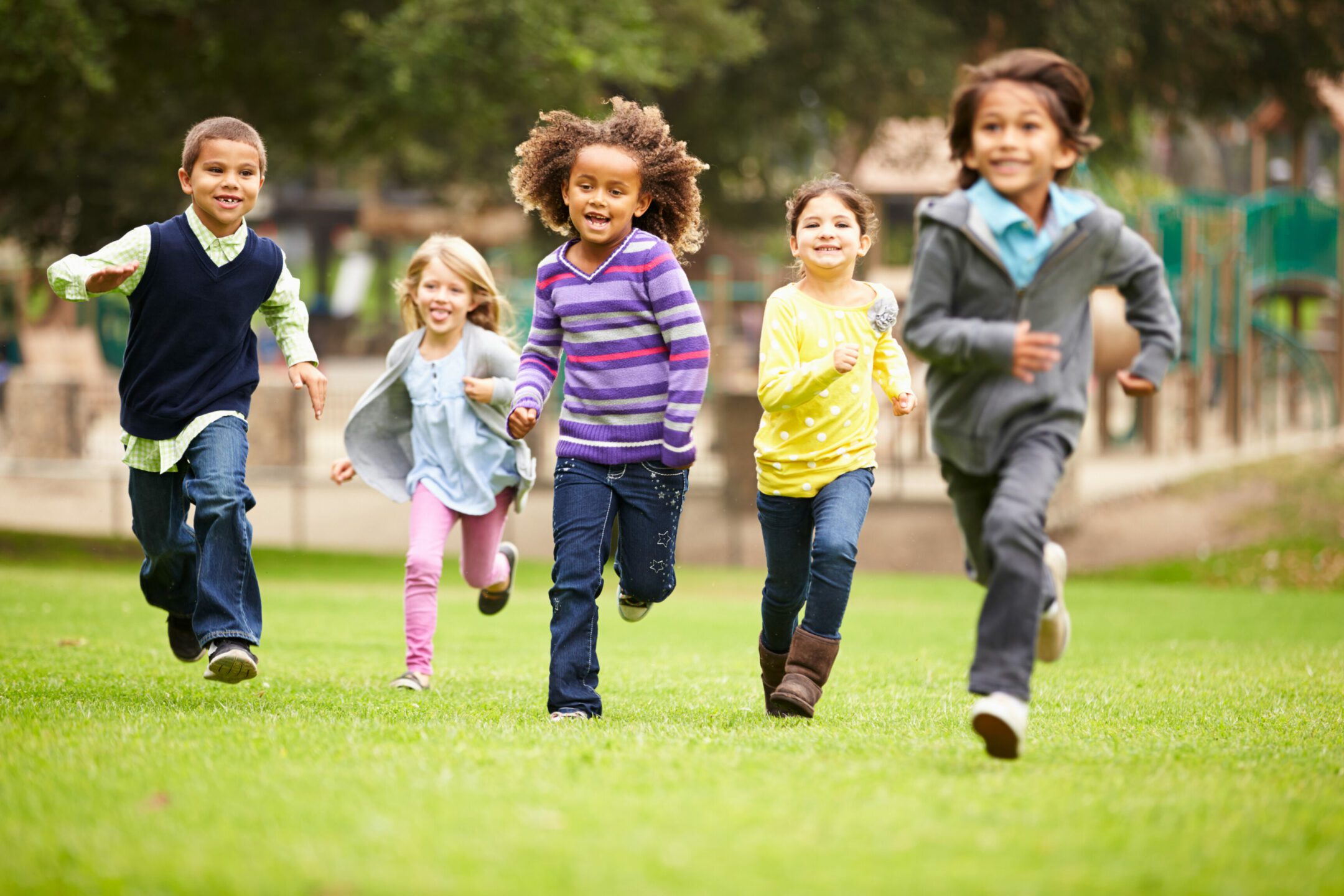 Smiling children running on grassy park field, playground behind.